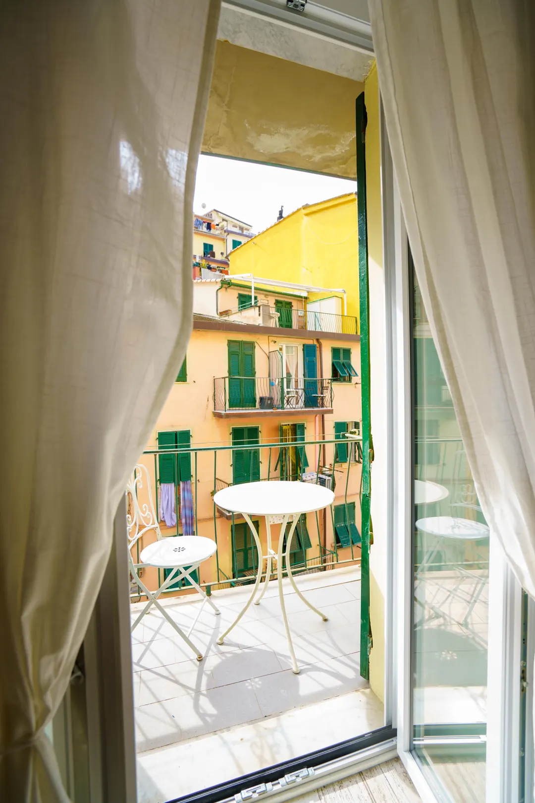 Balcone con tavolino bianco visto attraverso le tende, con le case colorate di fronte