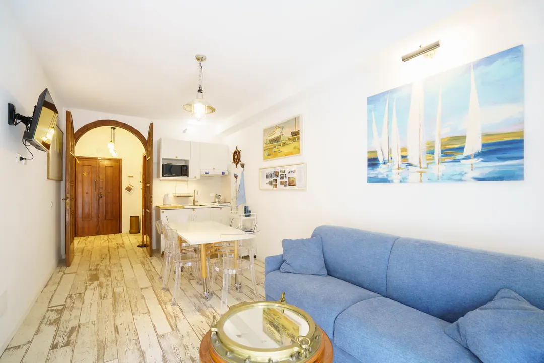 Soggiorno luminoso con divano azzurro, quadro di vele, tavolo bianco e cucina sullo sfondo